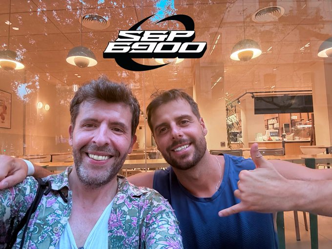 Two smiling men pose together inside a café, with the one on the right giving a shaka hand sign, beneath a stylized "SPX 6900" logo displayed at the top of the image.