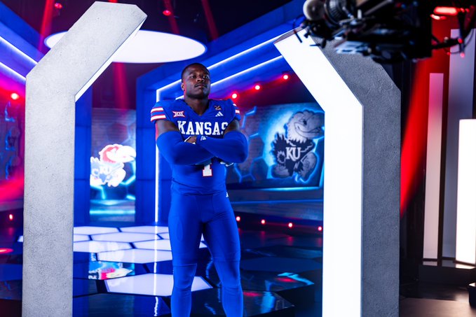 Kansas Jayhawks football player stands confidently with arms crossed in a blue uniform featuring the new "XRP" jersey patch, as he poses in a dramatic studio setting with neon blue and red lighting during Big 12 Media Day.