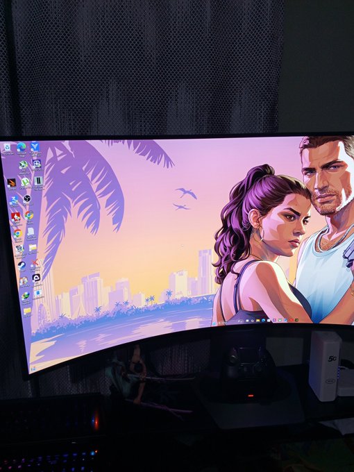 A curved monitor displaying a Grand Theft Auto-style wallpaper featuring two characters with a tropical sunset cityscape, with desktop icons visible on the left side.