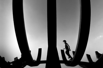 A black and white silhouette photograph showing a man walking across what appears to be a bridge or structure, framed by large curved architectural elements.