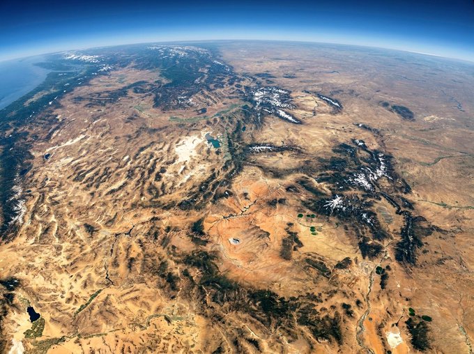 An aerial photograph taken from a high altitude shows a vast, arid landscape of mountains, valleys, and plateaus stretching to the horizon under a clear blue sky.