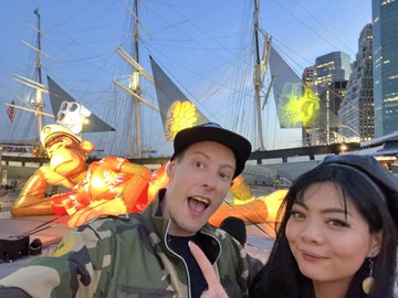 A selfie of a man and woman posing in front of a tall ship at a marina, with animated fire and ghost effects digitally added to the image.