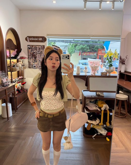 A young woman wearing a teddy bear ear headband, floral top, and green shorts takes a mirror selfie with her phone while puckering her lips, carrying a pink shoulder bag in a cozy boutique with a "Yafe" sign visible in the background. The cheerful morning selfie vibes perfectly match the "gm to those who still gm" crypto greeting, capturing that wholesome, feel-good energy to start the day.