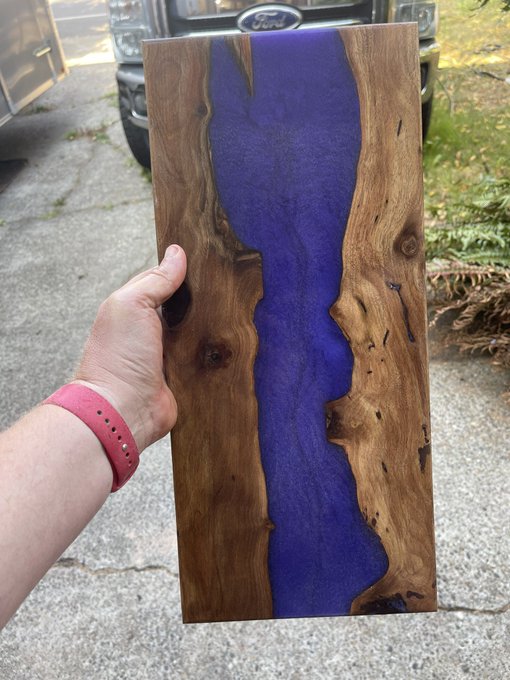 A hand wearing a pink watch band holds up a handmade wooden slab featuring a vibrant purple resin "river" flowing through the natural wood grain.