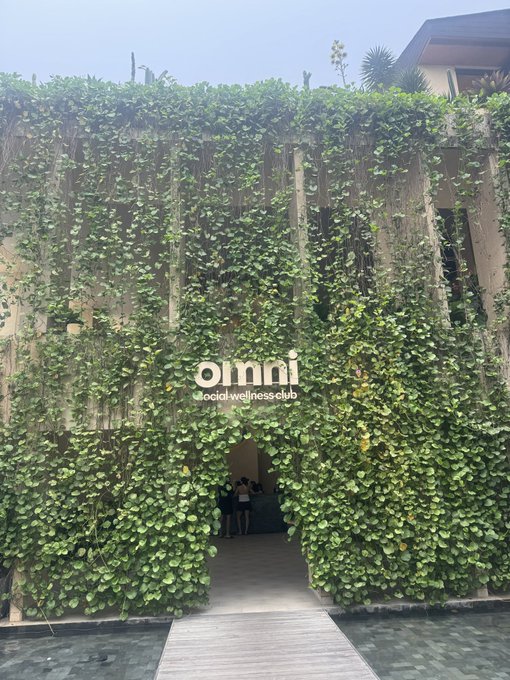 The image shows the lush, plant-covered entrance to "Omni social-wellness club," with the club's name displayed prominently amid cascading greenery. This aligns with the post's reference to "working at Omni," as the entrance serves as the physical location of the wellness club where the poster is based.