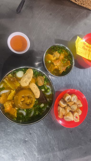 An overhead view of a Vietnamese meal featuring two bowls of soup with noodles and greens, a cup of tea, and a side of fried items on a metal table.