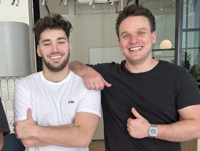 The image features two men standing close together, both smiling at the camera. They are wearing black t-shirts with white text that says "FTP" on them. The background of the photo includes a clock and a potted plant, adding some context to the scene. There is also an announcement about a partnership between Rainbet and streaming platform Kick.com, which seems to be confirmed by the image's content.
