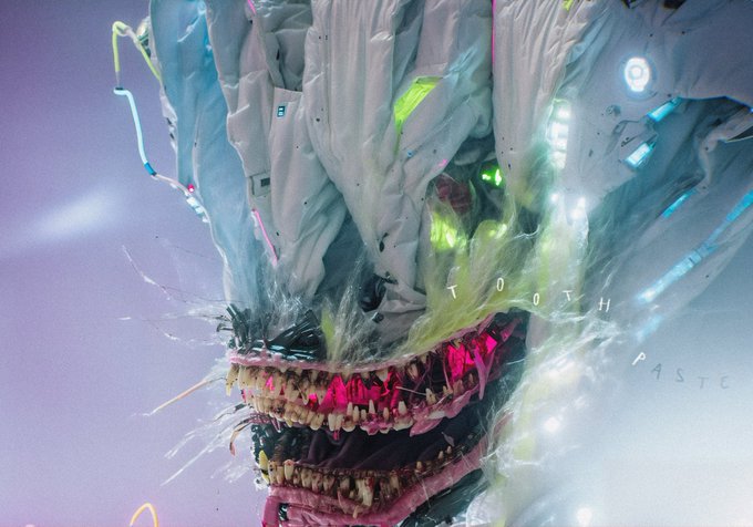 A surreal digital artwork depicts a monstrous figure made of melting fabric and plastic, with jagged teeth and glowing eyes, set against a pale background with faint text reading 'TOOTH PASTE'.