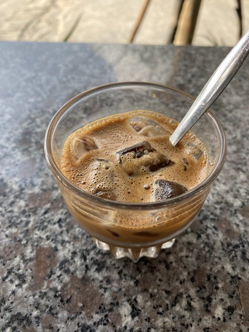 An iced coffee served in a clear glass with a metal straw, sitting on a granite countertop with sunlight filtering through.