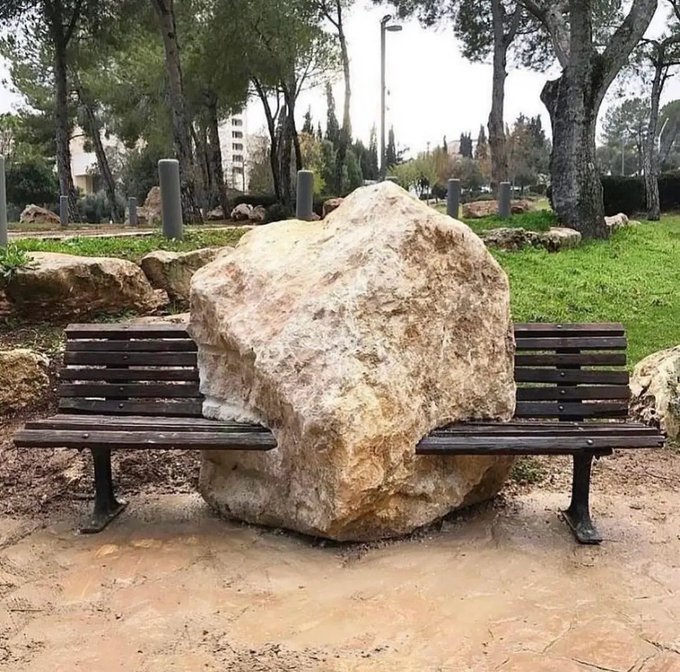 Two wooden park benches positioned on either side of a large boulder in an outdoor setting with trees and grass.