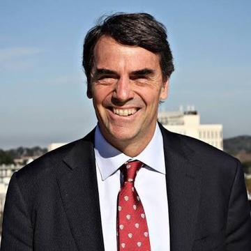 A smiling man in a dark suit with a red patterned tie stands outdoors against a backdrop of a clear blue sky and distant buildings, which aligns with the post about Tim Draper's bullish Bitcoin forecast. The image appears to be a portrait of the venture capitalist himself.