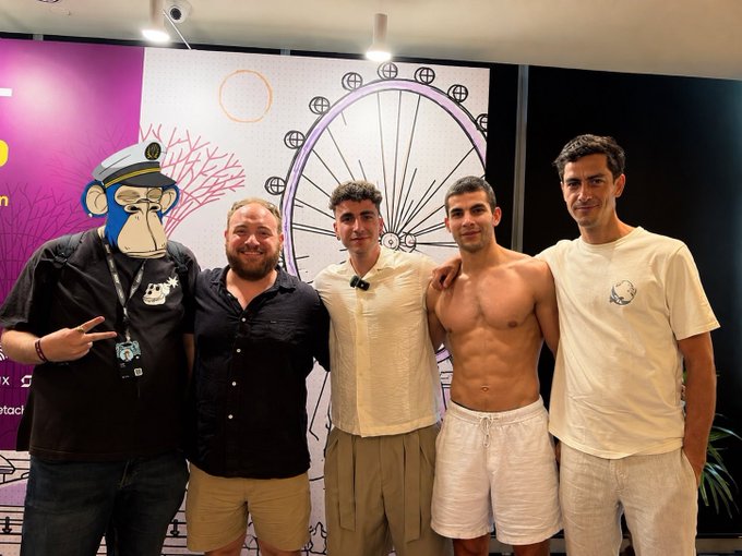 Five men pose together indoors at what appears to be an event, standing in front of a wall decorated with cartoon graphics including a monkey in a sailor hat.