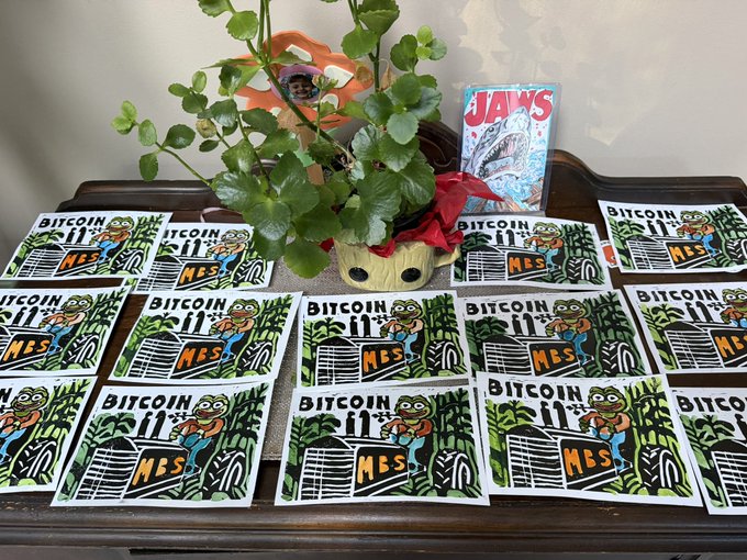 Multiple hand-printed and numbered art prints titled "BITCOIN MBS" featuring Pepe the Frog designs are arranged on a wooden table, with a potted plant and a "JAWS" DVD case displayed in the background, showcasing the physical collectible Bitcoin/Pepe artwork described in the post.
