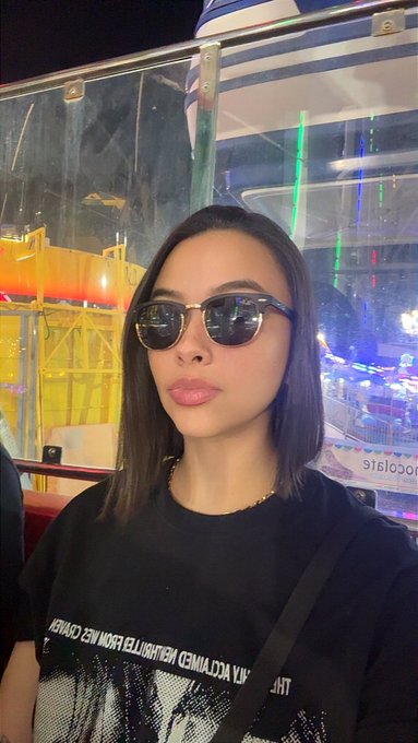 A selfie of a woman wearing dark sunglasses and a black graphic t-shirt, taken at night with blurred colorful lights and structures visible in the background.