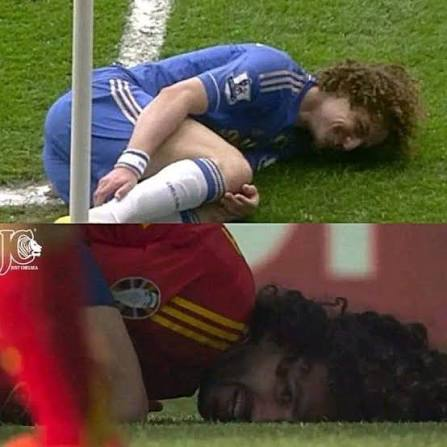 Two soccer players, one in a blue Chelsea jersey and another in a red and blue uniform, are shown lying on the grass field appearing to be injured during a match.