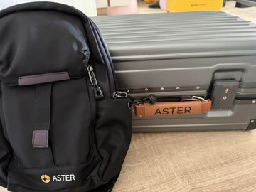 A black "ASTER" branded backpack rests against a dark hard-shell suitcase featuring a brown leather "ASTER" luggage tag, both items received as peripherals in July. The travel gear visually complements the post's theme of a spontaneous trip and appreciation for recent gifts from the Aster community.