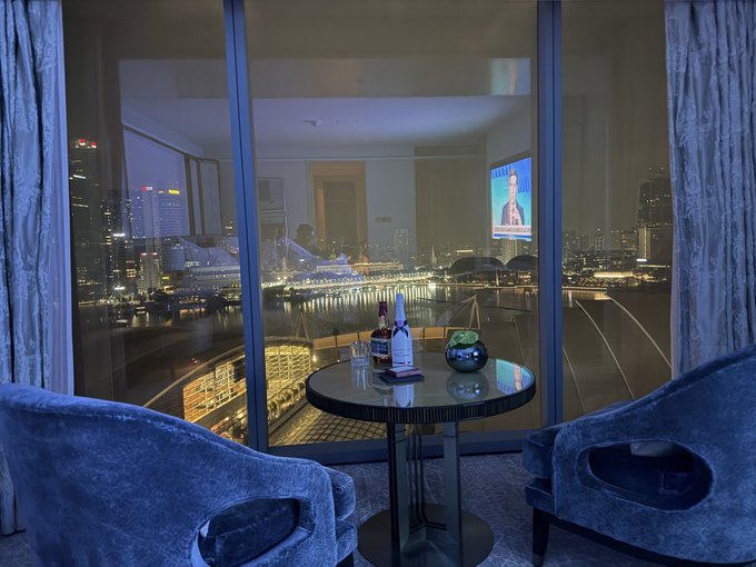 An evening view from inside a hotel room looking out a large window at a brightly lit cityscape and waterfront, with a small table holding bottles and glasses in the foreground.