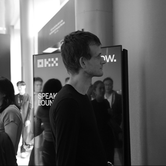 A man with short dark hair stands in profile at an event, wearing a dark t-shirt, with a display screen behind him showing "OKX" and "SPEAK LOUNGE" branding as attendees gather in the background.