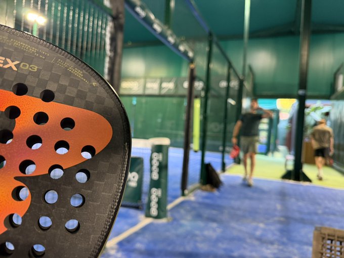 A padel racket marked "CX 03" dominates the foreground, while two players can be seen warming up on the court behind it, capturing the early morning padel session that contrasts with the post's late-night crypto auction news.