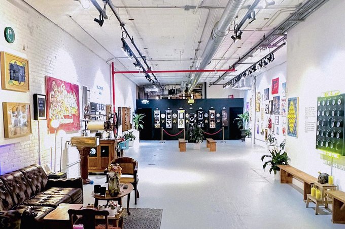 An industrial-style gallery space features exposed brick walls, track lighting, and polished concrete floors, with framed artworks and colorful pop-art pieces lining the walls, leather seating arrangements in the foreground, and display pedestals showcasing artwork in the center. The gallery is set up as a welcoming hub for art and Web3 community members to discover art and meet artists during NFTNYC week.