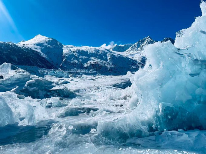 A glacier of jagged blue ice stretches beneath towering snow-capped mountains under a clear sky, symbolizing the frozen, treacherous landscape that altcoins must navigate. The image's harsh, melting terrain visually mirrors the post's warning that altcoins, unlike BNB and OKB, lack solid value support and could crash 50–70% even when the broader market only dips 20–30%.