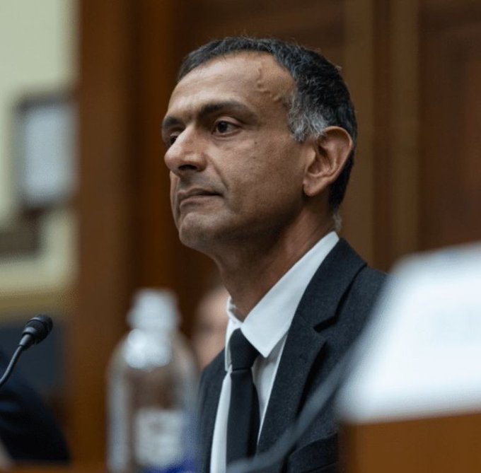 Portrait of a man in a dark suit with white shirt and dark tie, looking off to the side, with a microphone and water bottle visible, appearing to be in a formal hearing or courtroom setting.