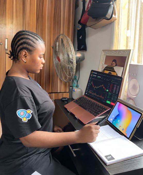 A young woman with braided hair working at a desk with a rose-gold laptop showing trading charts, a tablet, a notebook, and an electric fan beside her.