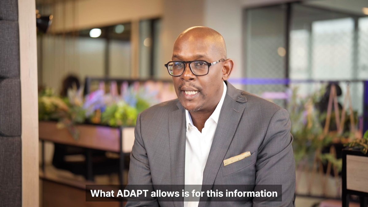The image shows a man wearing glasses sitting at a desk with his hands on his lap. He appears to be smiling for the camera while seated. The background of the image features a window, suggesting that he might be indoors or in an office setting. There is also text visible in the image, which reads "What AdaptPt allows is for this information". This implies that the man's suit and glasses are related to his work environment, possibly indicating a professional setting where he needs to adapt to different situations and communicate effectively with others.