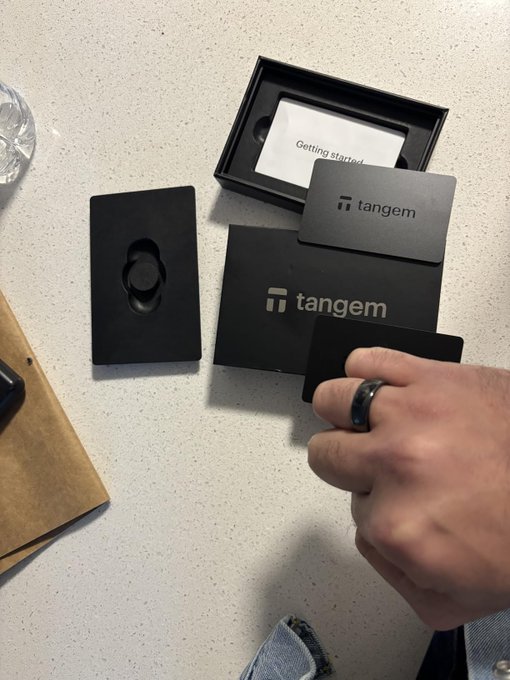 The image shows a person holding a black Tangem ring with their left hand. The ring has a unique design that resembles a ring but is actually a hardware wallet, specifically designed for storing private keys securely. The person's right hand is holding another black item, which appears to be a card or a piece of paper. The background features a white surface and some other objects such as a bottle and a book.