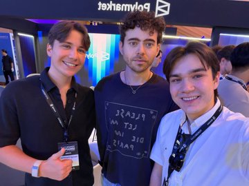 A photo of two young men (identified as Polygon co-founders Sandeep Nailwal and Mihailo Bjelic) posing with a young fan wearing a white shirt at what appears to be a Polygon event, with "Polygon" branding visible in the background.