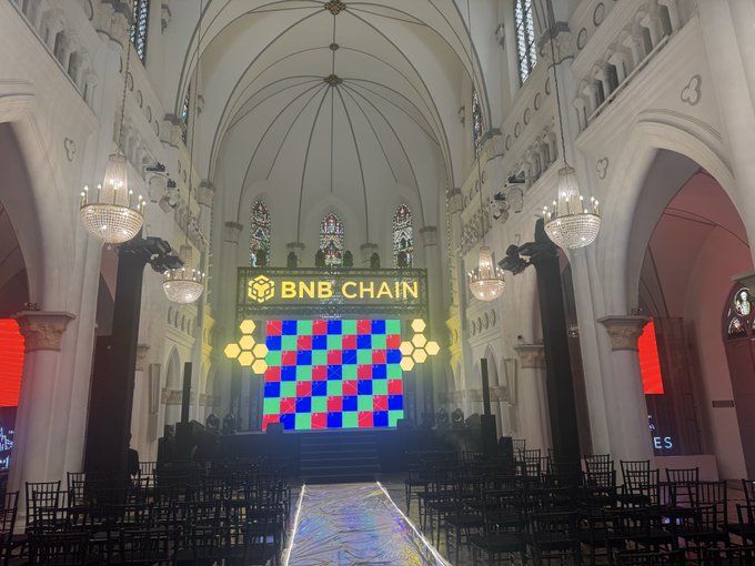 A grand cathedral interior with chandeliers and stained glass windows, with a 'BNB CHAIN' sign displayed on a colorful checkered screen at the altar.