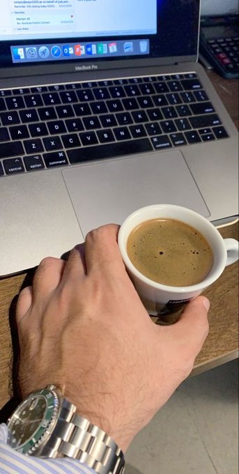A hand wearing a silver watch holds a small white espresso cup next to a MacBook Pro laptop displaying a webpage.