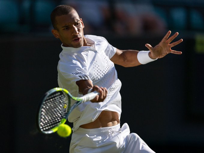 A focused tennis player in all-white attire, identified as Felix Auger-Aliassime, executes a powerful forehand swing at Wimbledon. The image relates to the post highlighting a $350,000 bet placed on him to defeat Novak Djokovic, with the payout tied to this high-stakes matchup.