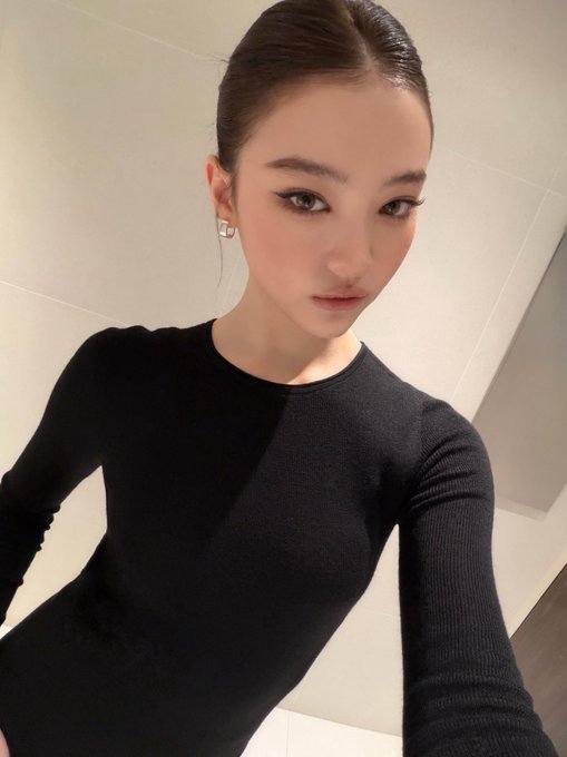 A young woman takes a high-angle selfie showcasing her sleek, tightly pulled-back hairstyle—the signature "slick bun"—paired with a fitted black long-sleeve top and defined makeup. Her polished, smooth hairline exemplifies the chic, controlled aesthetic referenced in the caption about the slick bun look.