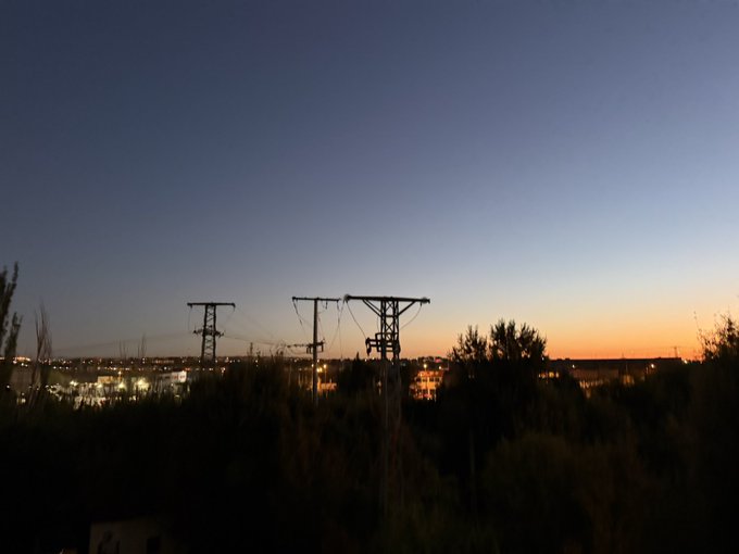 A twilight sky over a cityscape featuring silhouetted electrical transmission towers and power lines, with the horizon glowing in orange and the city lights beginning to illuminate below.