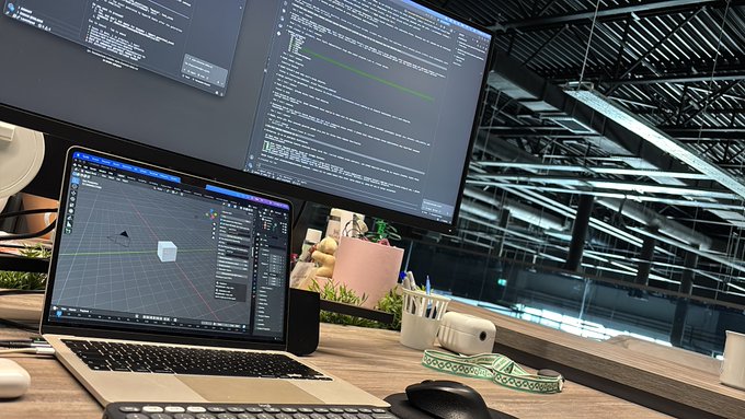A workspace featuring a laptop displaying 3D modeling software, alongside large monitors showing lines of code, in a room with an industrial glass ceiling.