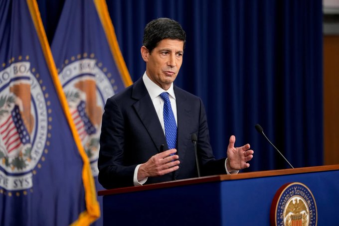A man in a dark suit and blue tie speaks at a podium with two microphones, standing before blue curtains and official-looking flags with partially visible seal text.