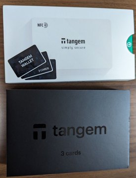 The image shows two wallet boxes, one black and one white, placed side by side on a wooden surface. The black box contains three cards while the white box has only two cards. Both boxes are labeled "Tagnem" with the word "Trust Wallet" written in black text. The post about the wallet boxes is titled "من افضل و حسن ال محافظ البارده حتي الان نسبه الامان عاليه و حاطه فيهم كل فلوسي الحمد الله ."