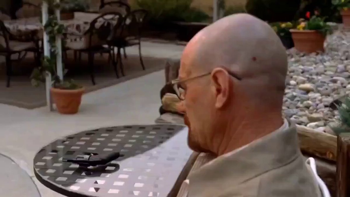 The image shows a bald man sitting at an outdoor table, wearing glasses and looking down. The scene takes place outdoors with potted plants around him. There are two chairs visible near the table where he's seated. A clock can be seen on one of the walls in the background. The post includes text that reads "POV: Told every person I've ever met in my life that XRP will have retire us all in 2026... Not even 6 months in to 2026:".