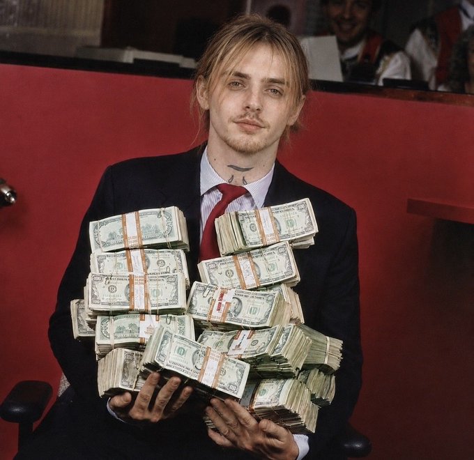 A man in a dark suit with a red tie holding an enormous stack of bundled U.S. hundred-dollar bills in his hands, posed against a red background.