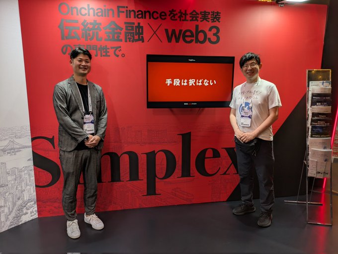 Two men stand smiling at the Simplex booth, which features bold red branding with Japanese text reading "Onchain Financeを社会実装 伝統金融×web3" and "手段は択ばない" (meaning "the means are not chosen" — any method is acceptable), promoting their JPYC EX system that bridges traditional finance with web3.