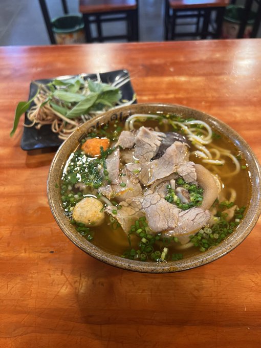 A hearty bowl of Asian noodle soup with sliced pork, mushrooms, and green onions in a clear broth, served alongside a small plate of greens on a wooden table.