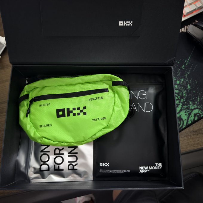 An opened OKX branded black box reveals a bright neon green fanny pack labeled "TRUSTED," "VERIFIED," and "SECURED" alongside the "24/7/365" text, sitting on top of packaging featuring the "DON'T FOR FUN" slogan and "THE NEW MONEY APP" tagline—showcasing the premium merchandise mentioned in the post.