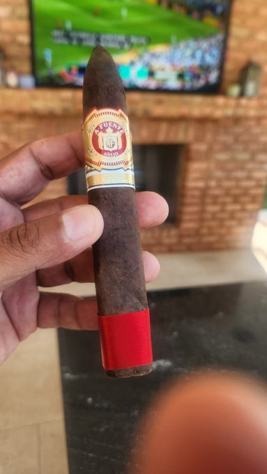A hand holding an 'Arturo Fuente' cigar in front of a TV showing a sports match in a living room with a brick fireplace.