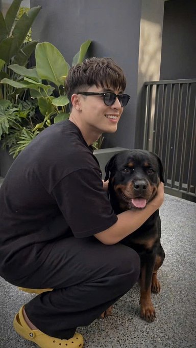 A photograph of a young man wearing sunglasses and a black t-shirt, crouching down and smiling while petting a Rottweiler dog outdoors next to a metal railing.