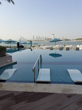 A serene morning view of an infinity pool overlooking a harbor with boats, lounge chairs with umbrellas, and a city skyline in the distance. The tranquil scene complements the post's message of finding peace and productivity in trading from any location, wishing followers a blessed and fruitful Friday.
