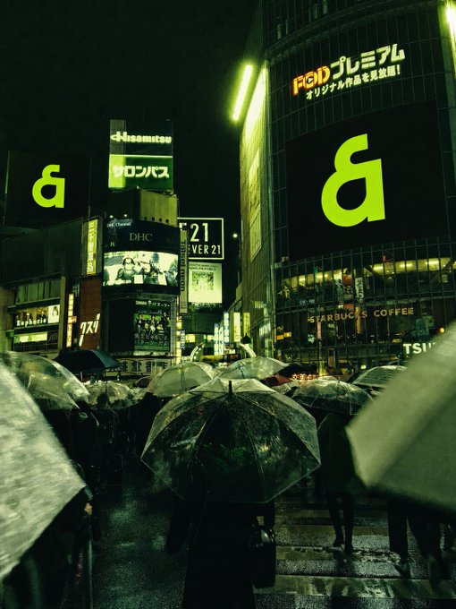 The image shows a group of people walking down a street at night, holding umbrellas over their heads as they navigate through the city. The scene takes place in front of tall buildings with neon signs lit up against the dark sky. A caption on the image reads "アジアのオンチェーン金融は、 @KaiaChain によって、よりシームレスに動き始めています". This phrase is likely a reference to the Kaia Chain, an online store that sells clothing and accessories.