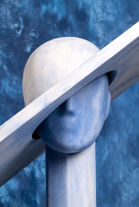 The image features a sculpture of a head with a hat that has been painted white. The hat appears to be tilted or upside down, giving the impression that the face is not visible. This artistic choice adds an interesting visual element to the piece. The sculpture is placed against a blue background, which further emphasizes its presence and draws attention to it.