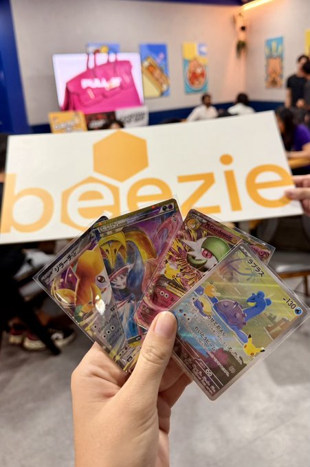 A hand holds up several shiny Japanese Pokémon TCG cards, including what appears to be a Gallade, Koffing, and a -100 HP Tangrowth, with the "Beezie" logo visible on a white sign in the background of what looks like a trading card event. The post's caption about "crazy pulls at the @Beezie event with my frens" matches the celebratory display of the valuable holographic cards fanned out for the camera.