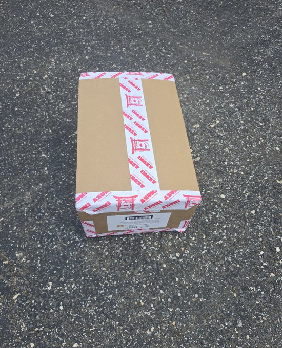 A sealed cardboard box with pink and white "Azuki" branded tape sits on asphalt, addressed to a buyer as part of an "Alley Runner Pre-sale" from Azuki, suggesting the package contains a pre-sale trading card or NFT-related merchandise that has been kept sealed.